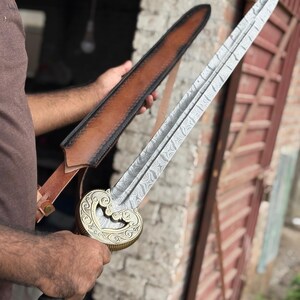 May include: A detailed image of a decorative sword with a patterned blade and a gold-colored hilt. The sword is held near a brown leather sheath. The hilt features an ornate design, and the blade has a distinctive pattern.