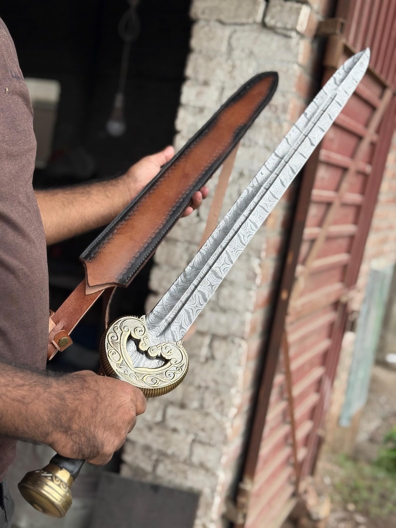 May include: A detailed image of a decorative sword with a patterned blade and a gold-colored hilt. The sword is held near a brown leather sheath. The hilt features an ornate design, and the blade has a distinctive pattern.
