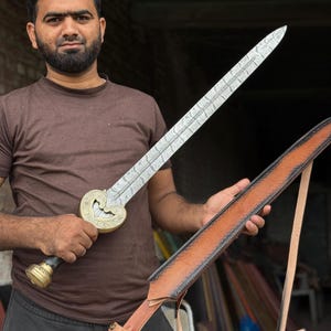 May include: A decorative sword with a silver blade and a gold-colored hilt. The sword has an ornate design and is held with a brown leather sheath. The sword's blade has a length of approximately 24 inches.
