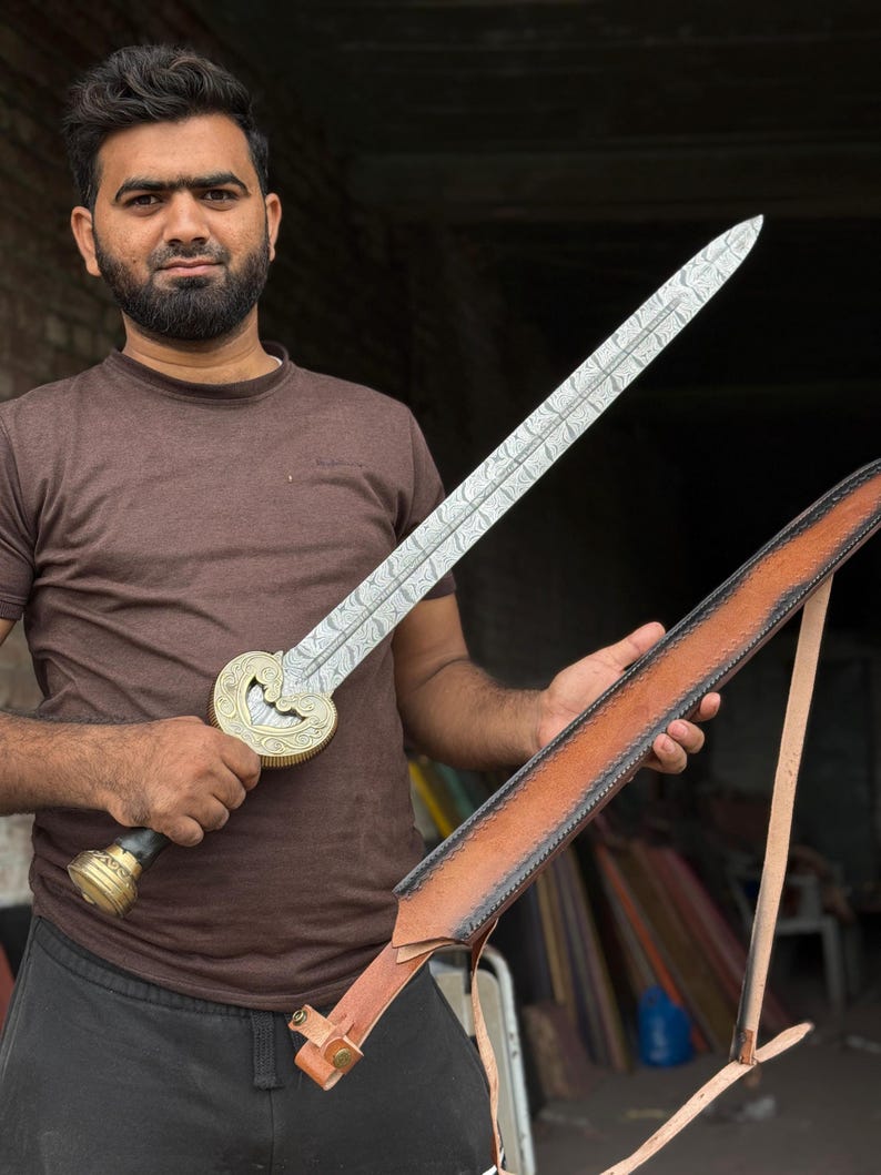 May include: A decorative sword with a silver blade and a gold-colored hilt. The sword has an ornate design and is held with a brown leather sheath. The sword's blade has a length of approximately 24 inches.