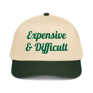Puede incluir: Gorra de béisbol con corona color crema y visera verde oscuro. La gorra presenta el texto "Expensive & Difficult" en escritura verde en la parte delantera. La gorra tiene un botón verde oscuro en la parte superior.