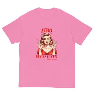 Puede incluir: Una camiseta rosa con una ilustración retro de una mujer con un vestido rojo haciendo el gesto de la mano "ok". El texto dice "ZERO FUCKS GIVEN" en rojo, con "Butch Unicorn" debajo. Es una prenda informal.