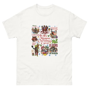 May include: White t-shirt with a vibrant graphic from "The Muppet Christmas Carol." The design incorporates characters, text, and festive imagery. Text includes "You Find Love," "Light the lamps," and "No Cheeses."