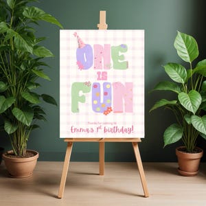 Op de afbeelding: Een kleurrijk verjaardagsbord op een ezel met de tekst "ONE IS FUN". Het bord heeft een roze en witte geruite achtergrond en de tekst "Bedankt voor het komen naar Emma's 1e verjaardag!" is ook opgenomen.