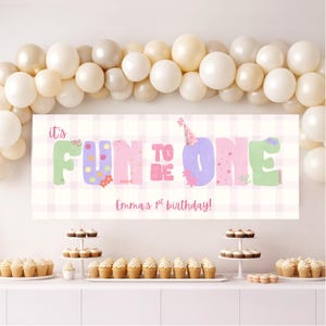 Puede incluir: Una pancarta colorida con la frase "It's FUN TO BE ONE", adornada con flores y lunares, se exhibe en una fiesta de cumpleaños. Globos y cupcakes completan la decoración festiva.