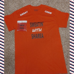 Puede incluir: Camiseta naranja de manga corta con el texto "SWEATIN' WITH SHAREA" impreso en la parte delantera. La camiseta tiene "SWEAT" impreso en la manga izquierda y "CREW" impreso en la manga derecha. Una pequeña etiqueta roja con el texto "SWEAT CREW" está unida a la parte delantera de la camiseta.