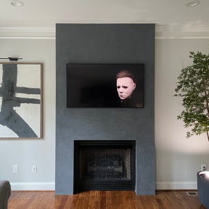 May include: A gray fireplace with a black mantel and a black screen in front of the fireplace. A flat screen TV is mounted above the fireplace. A black and white abstract painting is on the wall to the left of the fireplace. A potted plant is on the right side of the fireplace.