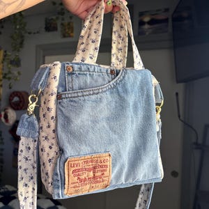 May include: A handmade tote bag crafted from light blue denim fabric and cream-colored floral print fabric. The bag features a front denim pocket, a Levi Strauss & Co. leather patch, and a detachable shoulder strap with gold-tone metal hardware.