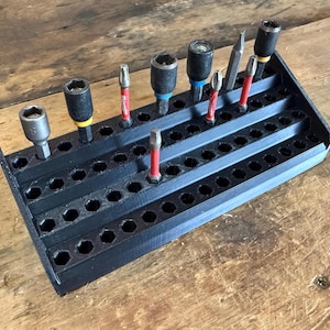 May include: A black tool organiser with multiple rows of hexagonal holes, holding various screwdriver bits and sockets. The tools have red, yellow, and blue accents. The organiser is made of plastic and sits on a wooden surface.