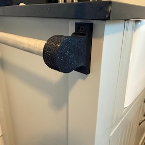 Könnte beinhalten: Ein schwarzer, verzierter Handtuchhalter mit einem dekorativen Endstück ist an einem weißen Schrank befestigt. Die Stange besteht aus einem hellen Material. Der Schrank hat eine dunkle Arbeitsplatte und ein weißes Waschbecken.