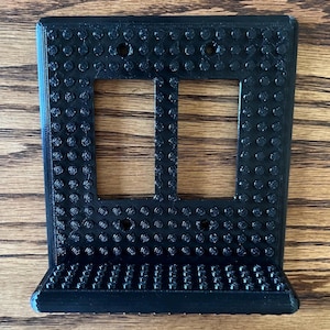 Puede incluir: Cubierta de interruptor de luz doble negra y texturizada con un estante. La cubierta tiene una cuadrícula de pequeños círculos en relieve y dos aberturas rectangulares para los interruptores. El estante está en la parte inferior.