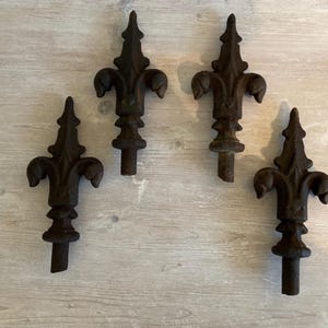 May include: Four dark brown fleur-de-lis finials, each with a pointed top and three curved petals. The finials have a textured, aged appearance and are arranged on a light wood-grain surface.