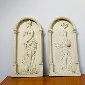 Op de afbeelding: Een paar witte gipsen wandsculpturen met twee vrouwen in klassieke poses. Elk beeld is omlijst door een decoratieve boog en kolommen.