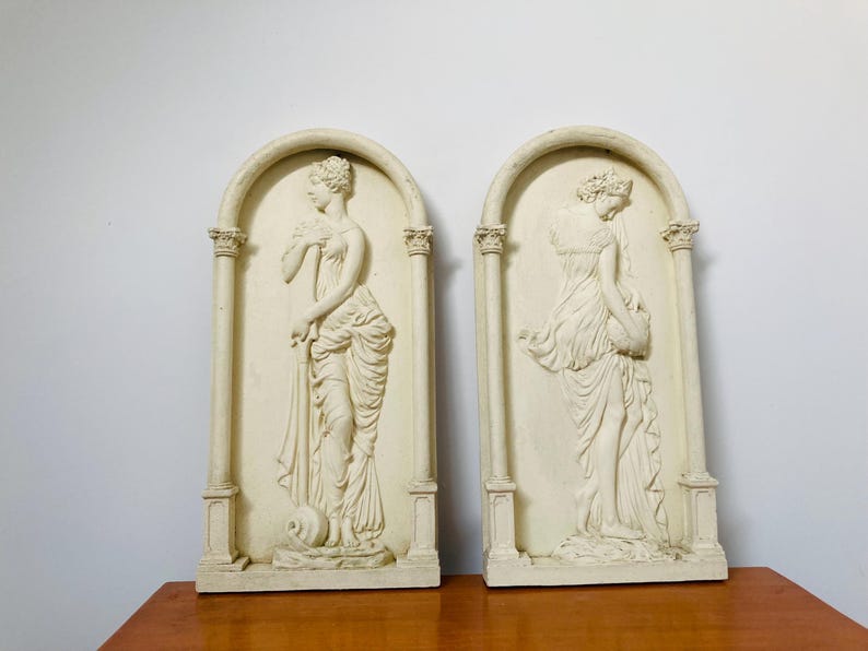 Op de afbeelding: Een paar witte gipsen wandsculpturen met twee vrouwen in klassieke poses. Elk beeld is omlijst door een decoratieve boog en kolommen.