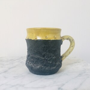 May include: A ceramic mug with a yellow top and a black textured bottom. The mug has a handle and is sitting on a white marble surface.