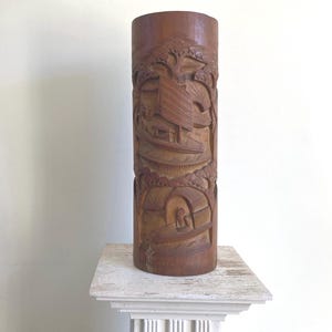 May include: A carved wooden cylinder vase featuring a detailed design of a boat and trees. The vase is brown and has a natural wood finish.