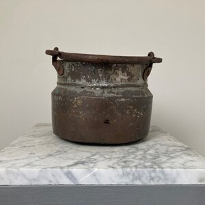 May include: An antique copper pot with two iron handles. The pot has a worn, aged patina and is sitting on a white marble surface.