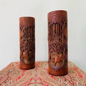 May include: Two tall, cylindrical, brown bamboo vases with intricate carvings of trees and animals. The vases are on a red and gold paisley patterned fabric.