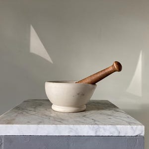 May include: A cream-coloured mortar and pestle set. The mortar is a rounded bowl with a small base, and the pestle is a cylindrical shape with a rounded head. The set is made of stone and wood. The mortar and pestle are on a marble surface.