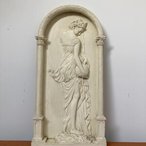 Op de afbeelding: Een witte gipsen wandsculptuur van een vrouw in een vloeiende jurk, staand in een deuropening met kolommen aan weerszijden. De sculptuur is in reli&euml;fstijl, waarbij de vorm van de vrouw uit de achtergrond steekt.