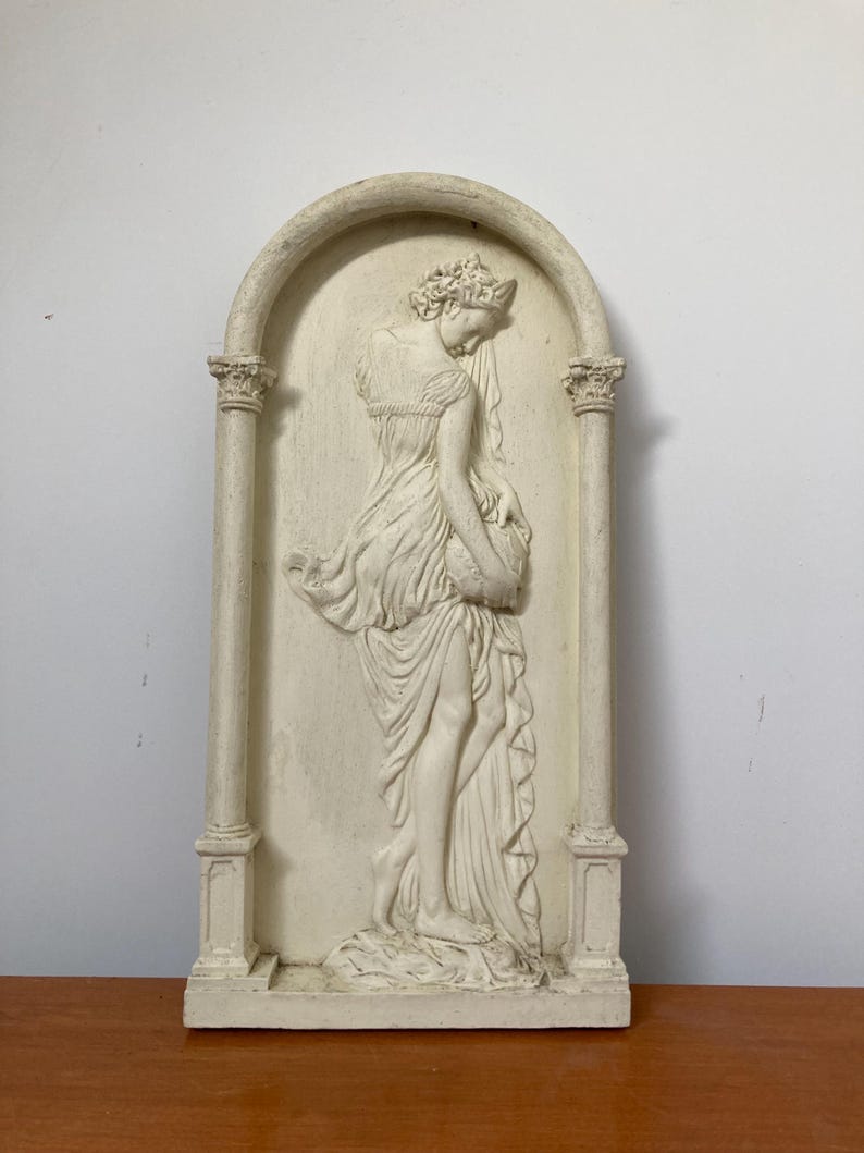 Op de afbeelding: Een witte gipsen wandsculptuur van een vrouw in een vloeiende jurk, staand in een deuropening met kolommen aan weerszijden. De sculptuur is in reli&euml;fstijl, waarbij de vorm van de vrouw uit de achtergrond steekt.