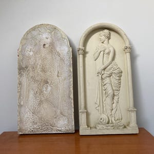 Op de afbeelding: Twee witte wandplaquettes van gips, een met een reli&euml;f van een vrouw in een vloeiende jurk, de andere met een textuur.