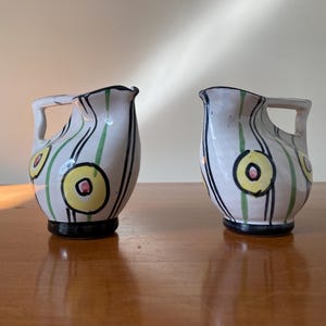 May include: Two white ceramic jugs with green and black stripes and yellow and pink circles. The jugs have a handle on the side.