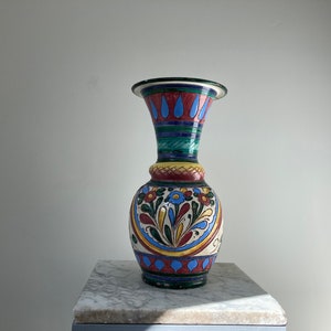 May include: A decorative ceramic vase with a flared top and a bulbous base. The vase has a colourful floral and geometric design in red, blue, yellow, and green. The vase is approximately 30 cm tall.