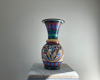 May include: A tall, decorative ceramic vase with a flared top and a bulbous base. The vase features a colorful floral and geometric design in red, blue, yellow, and green. The vase is approximately 12 inches tall.