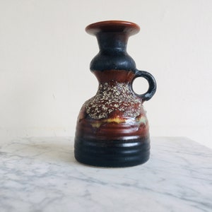 May include: A brown and black ceramic candle holder with a textured surface and a handle. The holder has a wide base and a narrow neck.