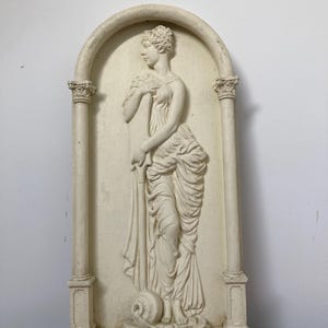 Op de afbeelding: Een witte gipsen wandsculptuur van een vrouw in een vloeiende jurk, staand in een decoratieve boog met kolommen. De sculptuur is in reli&euml;fstijl, met de vorm van de vrouw licht verhoogd ten opzichte van de achtergrond.