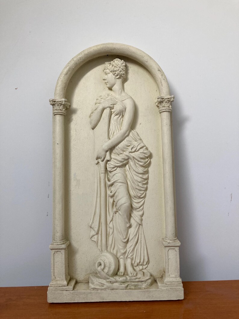 Op de afbeelding: Een witte gipsen wandsculptuur van een vrouw in een vloeiende jurk, staand in een decoratieve boog met kolommen. De sculptuur is in reli&euml;fstijl, met de vorm van de vrouw licht verhoogd ten opzichte van de achtergrond.