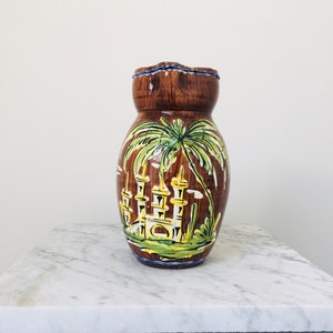 May include: A brown ceramic jug with a hand-painted design of a palm tree and a castle. The jug has a scalloped rim and a brown glaze.