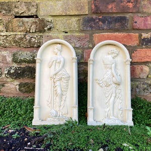 Op de afbeelding: Twee witte gipsen wandplaquettes met reli&euml;f sculpturen van vrouwen in vloeiende gewaden. De plaquettes zijn rechthoekig met een afgeronde boven- en onderkant en hebben een witte achtergrond.