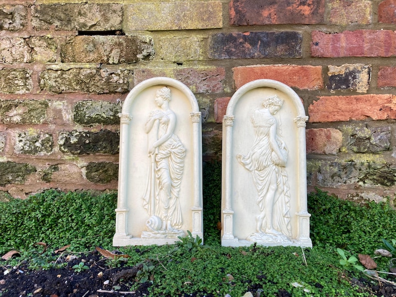 Op de afbeelding: Twee witte gipsen wandplaquettes met reli&euml;f sculpturen van vrouwen in vloeiende gewaden. De plaquettes zijn rechthoekig met een afgeronde boven- en onderkant en hebben een witte achtergrond.