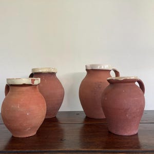 May include: A collection of four terracotta pitchers with rounded bodies and handles. Each vessel features a distinct matte reddish-brown clay finish and a contrasting smooth, off-white glazed rim around the top opening of the container.