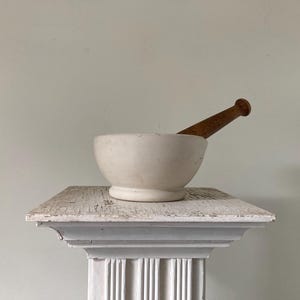 May include: A white mortar and pestle set with a wooden pestle, resting on a white, distressed pedestal. The mortar is a simple, round bowl shape. The pestle is brown and angled across the bowl.