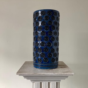 May include: A tall, blue ceramic vase with a repeating pattern of black circles. The vase is sitting on a white pedestal.