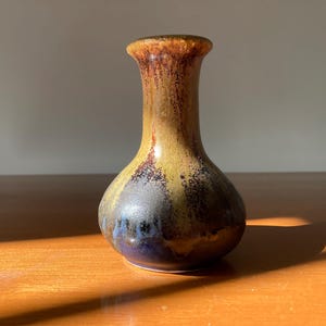 May include: A small, ceramic vase with a rounded body and a narrow neck. The vase is decorated with a mottled glaze in shades of brown, yellow, and blue.