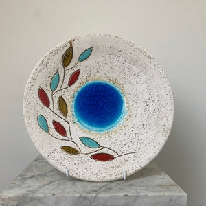 May include: A white ceramic plate with a blue centre and a stylised leaf design in red, brown, and blue.