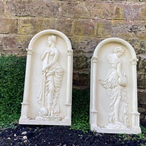 Op de afbeelding: Twee witte gipsen wandplaquettes met reli&euml;fsculturen van vrouwen in vloeiende gewaden. De plaquettes zijn rechthoekig met een afgeronde bovenkant en hebben een witte achtergrond.
