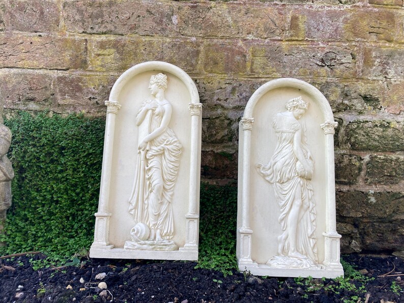 Op de afbeelding: Twee witte gipsen wandplaquettes met reli&euml;fsculturen van vrouwen in vloeiende gewaden. De plaquettes zijn rechthoekig met een afgeronde bovenkant en hebben een witte achtergrond.