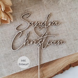 Könnte beinhalten: Ein Kuchenaufsatz aus Holz mit den Namen "Sandra" und "Christian" in eleganter Schreibschrift, verbunden durch ein Herzsymbol. Der Aufsatz ist an einem dünnen Stab befestigt. Der Text "Inkl. Entwurf" ist ebenfalls sichtbar.