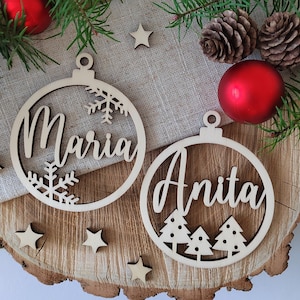 K&ouml;nnte beinhalten: Zwei h&ouml;lzerne Weihnachtsbaumschmuckst&uuml;cke mit den ausgeschnittenen Namen "Maria" und "Anita". Die Schmuckst&uuml;cke sind mit Schneeflocken und Tannenb&auml;umen verziert.