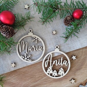 K&ouml;nnte beinhalten: Zwei h&ouml;lzerne Weihnachtsbaumschmuckst&uuml;cke mit den Namen "Maria" und "Anita" darauf. Das Ornament mit "Maria" hat Schneeflocken, und das Ornament mit "Anita" hat Weihnachtsb&auml;ume.