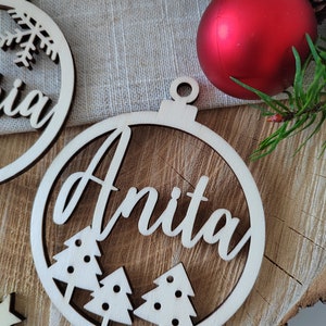 K&ouml;nnte beinhalten: Ein h&ouml;lzerner Weihnachtsbaumschmuck mit dem Namen "Anita" in Kursivschrift. Der Schmuck ist kreisf&ouml;rmig mit einer kleinen Schlaufe oben zum Aufh&auml;ngen. Drei kleine Weihnachtsb&auml;ume sind aus dem Design des Schmucks ausgeschnitten.