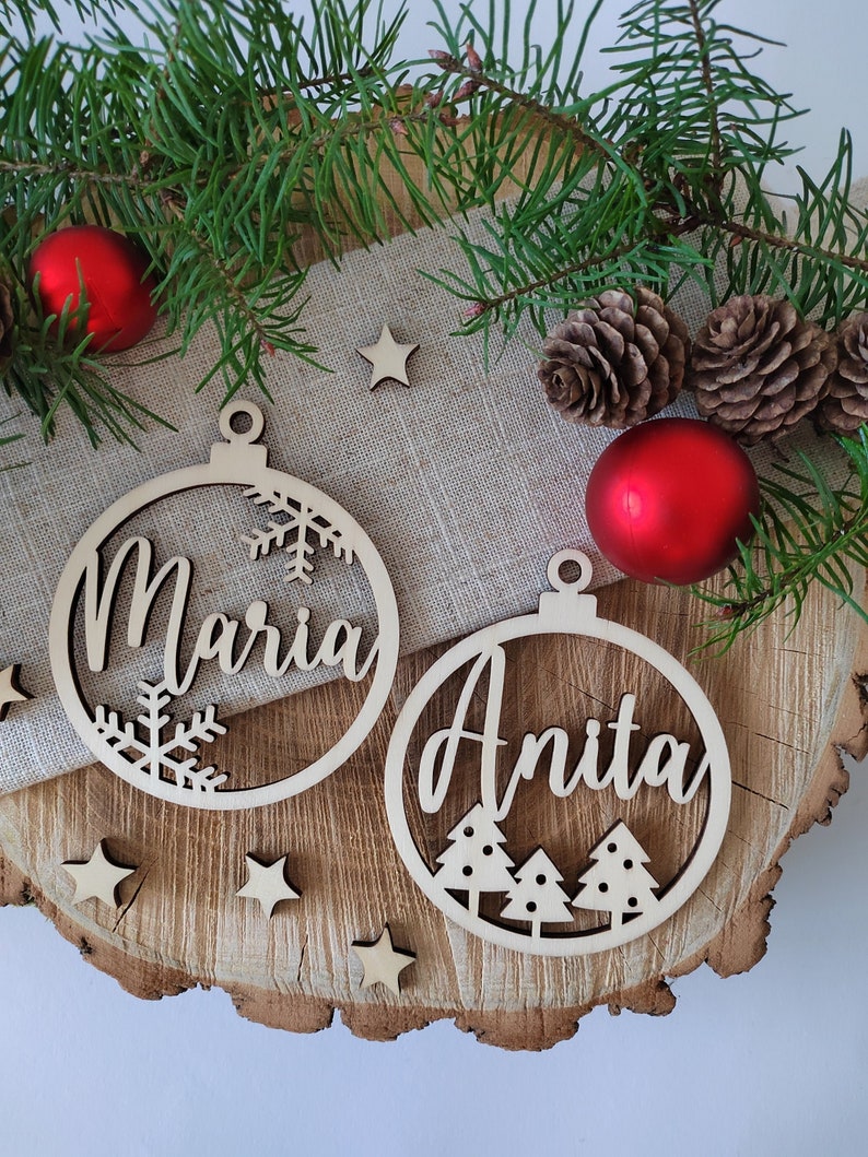 K&ouml;nnte beinhalten: Zwei h&ouml;lzerne Weihnachtsbaumschmuckst&uuml;cke mit den ausgeschnittenen Namen "Maria" und "Anita". Die Schmuckst&uuml;cke sind mit Schneeflocken und Tannenb&auml;umen verziert.