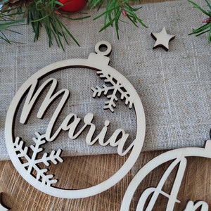 K&ouml;nnte beinhalten: Zwei h&ouml;lzerne Weihnachtsbaumschmuckst&uuml;cke mit den Namen "Maria" und "Ann" ausgeschnitten in der Mitte. Die Schmuckst&uuml;cke sind mit Schneeflocken verziert und haben ein Loch oben zum Aufh&auml;ngen.