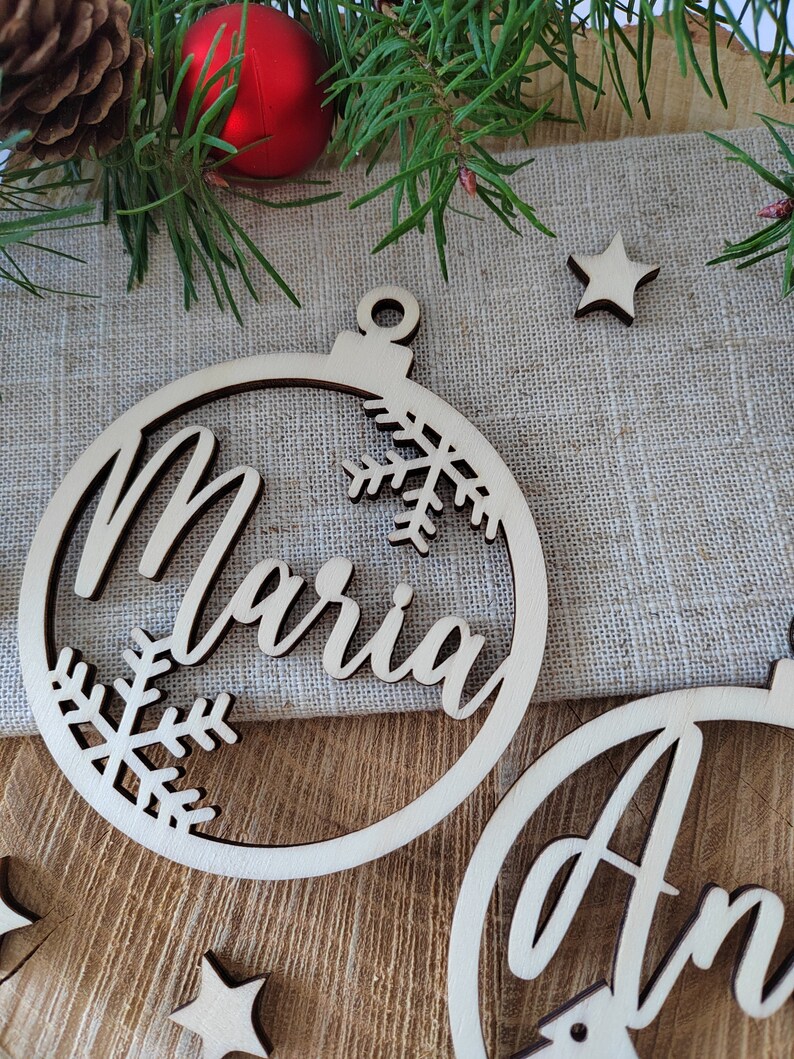 K&ouml;nnte beinhalten: Zwei h&ouml;lzerne Weihnachtsbaumschmuckst&uuml;cke mit den Namen "Maria" und "Ann" ausgeschnitten in der Mitte. Die Schmuckst&uuml;cke sind mit Schneeflocken verziert und haben ein Loch oben zum Aufh&auml;ngen.
