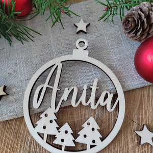 K&ouml;nnte beinhalten: Ein runder Weihnachtsbaumschmuck aus Holz mit dem ausgeschnittenen Namen "Anita" in der Mitte. Der Schmuck ist mit drei kleinen Weihnachtsb&auml;umen verziert und hat oben ein Loch zum Aufh&auml;ngen.